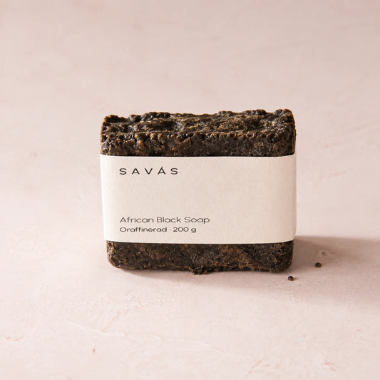 En bit svart tvål i sitt pappersband med SAVÁS tryckt på etiketten, på ljus rosa bakgrund