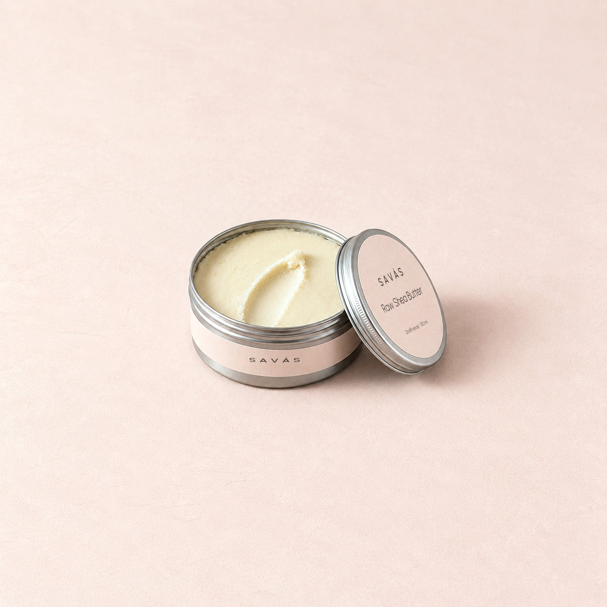 En öppen aluminiumburk med Raw Shea Butter från SAVÁS, locket lutat mot burken, på rosa bakgrund